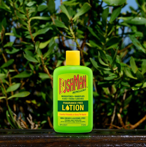 A bottle of Bushman Repellent Moisturized Tick and Bug Repellent Fragrance Free Lotion, placed on a wet wooden surface with a blurred background of green foliage and blue sky.